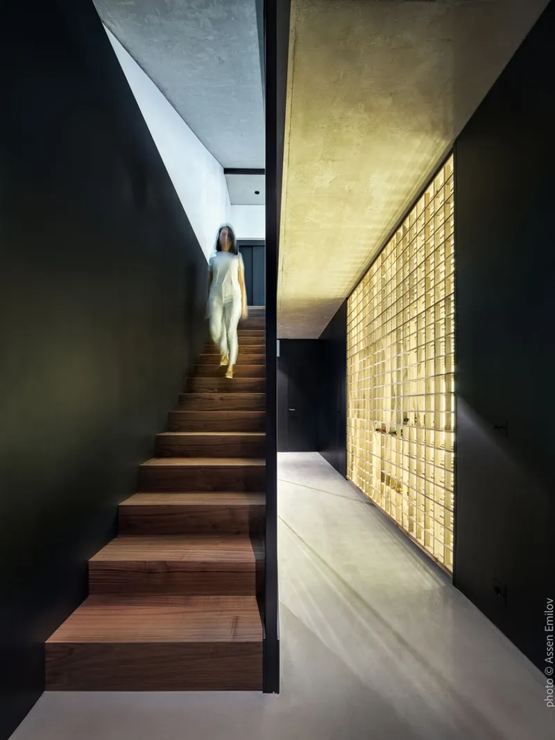 Timber staircase rising between dark walls alongside a backlit glass block wall with figure ascending