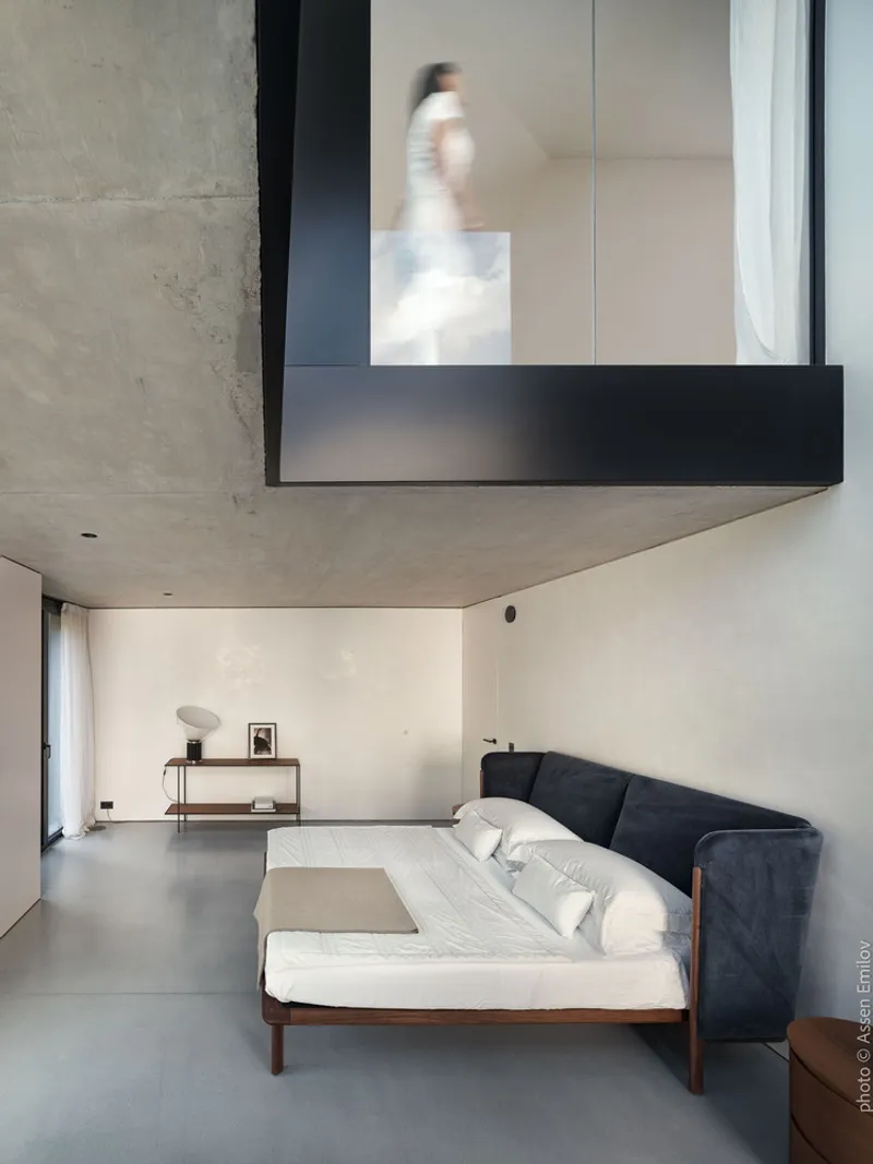 Double-height bedroom with cantilevered black-framed window box and blurred figure moving above