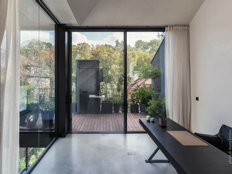 Interior living space opening onto a timber deck terrace with potted plants and autumn trees beyond