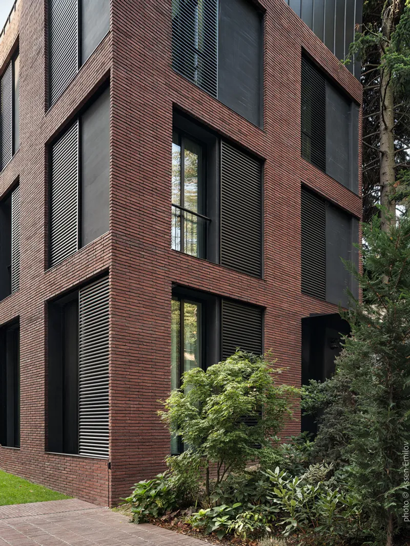 Brick facade with integrated horizontal louvered screens beside mature evergreen trees and landscaped plantings