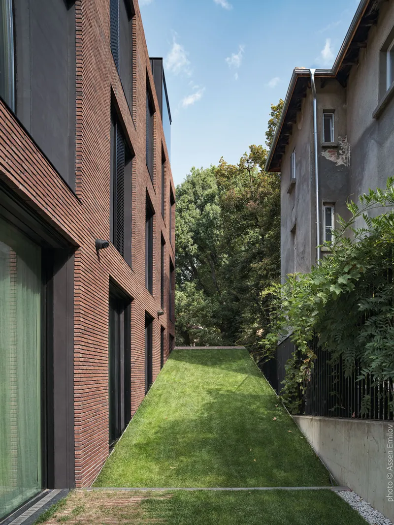Grass lawn corridor between new brick volume and adjacent stucco house under clear blue sky
