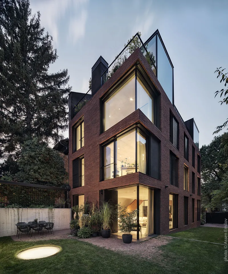 Corner volume with illuminated floor-to-ceiling windows at dusk beside paved terrace and potted grasses