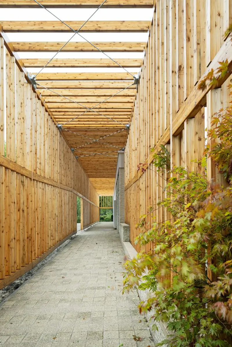 Covered timber walkway with vertical wood slatted walls and planted foliage along the gravel path