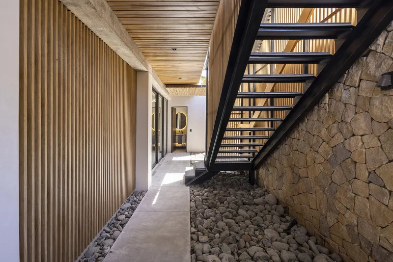 Exterior passageway with vertical timber screening and open-tread steel stair beside a stone wall and gravel bed