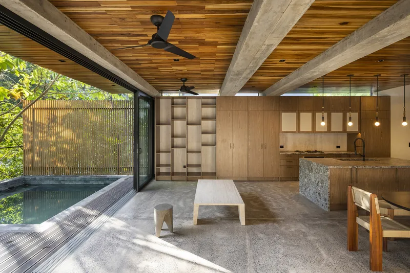 Open-plan living space with timber ceiling beams, concrete floor, and sliding glass doors opening to a plunge pool and slatted timber screen