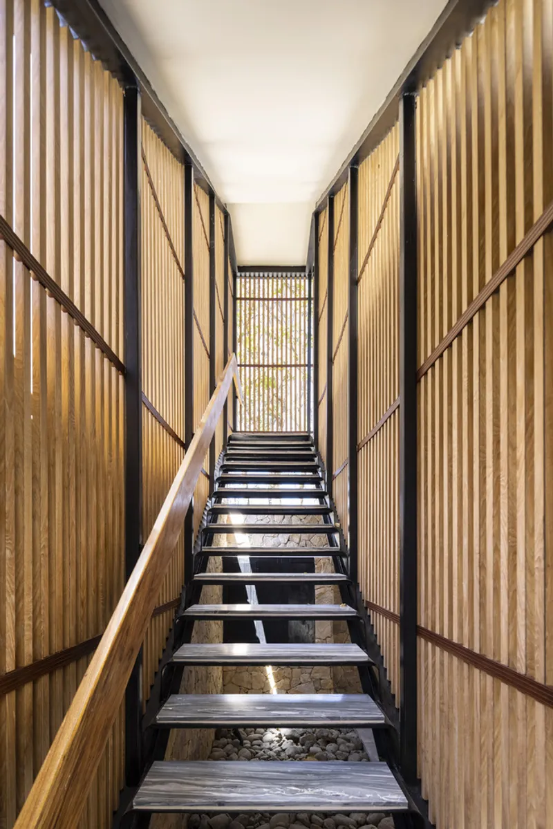Metal staircase with open risers flanked by vertical timber slat walls and wooden handrails, with dappled sunlight filtering through from above