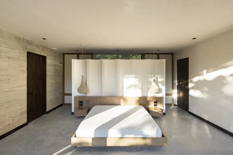 Bedroom with polished concrete floor, timber platform bed, and white paneled headboard wall with pendant lights casting shadows in afternoon sun