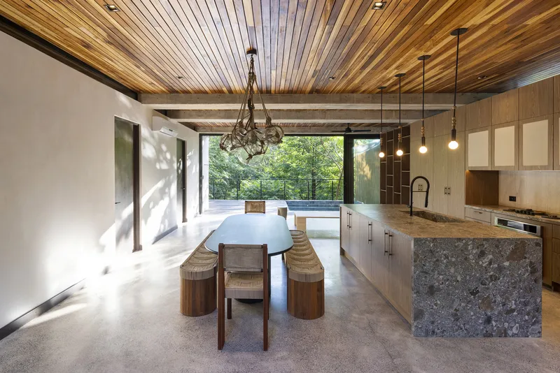 Open-plan dining and kitchen with striped timber ceiling and pendant lights framing views to the forest