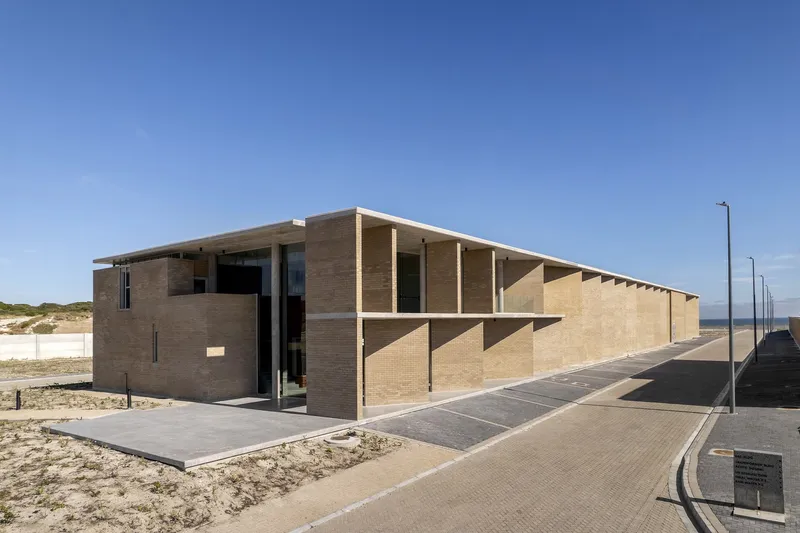 Buff brick volume with deep columned portico beside coastal dunes and paved road