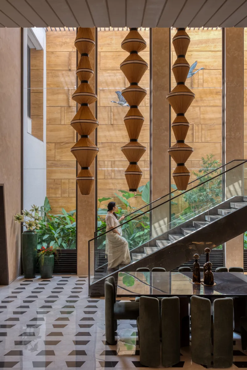 Steel staircase with glass railing ascending past stacked wooden pendant lights and planted courtyard below