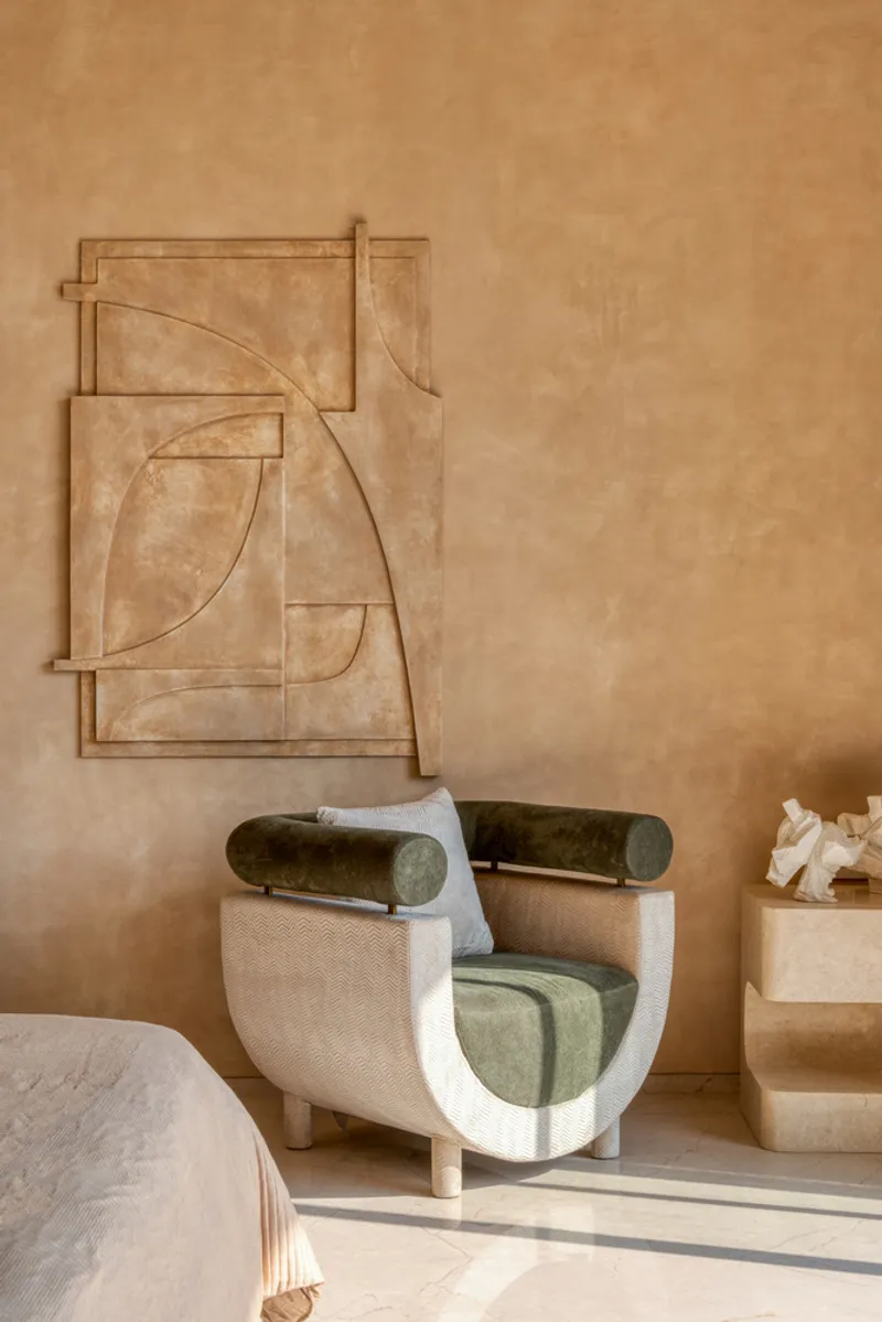 Carved stone relief panel with geometric curves mounted above a curved upholstered armchair in morning sunlight