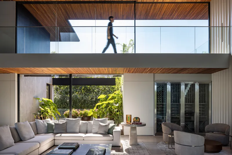 Double-height living room with timber ceiling and glass bridge above as a figure crosses