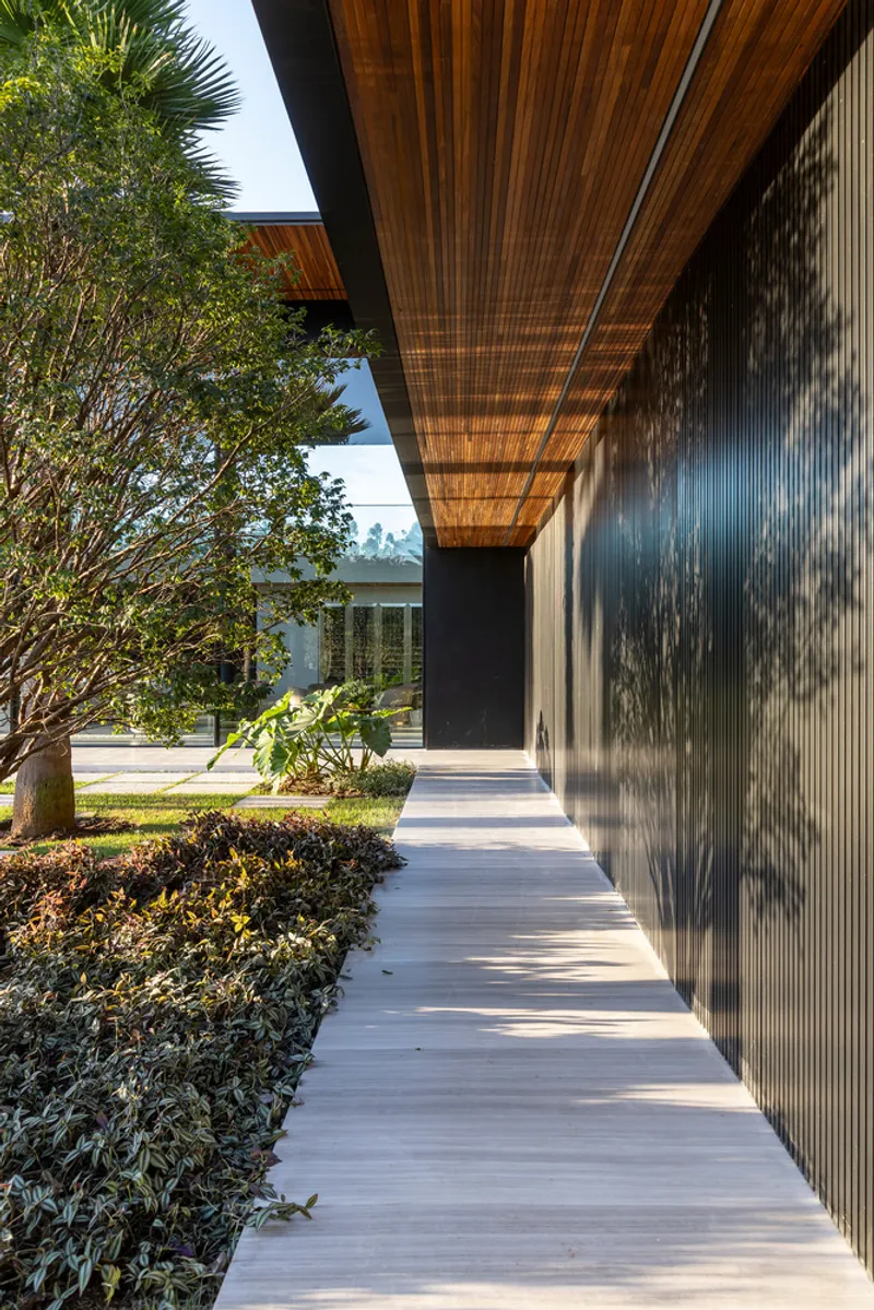 Covered concrete pathway alongside corrugated metal wall with dappled tree shadows under timber canopy