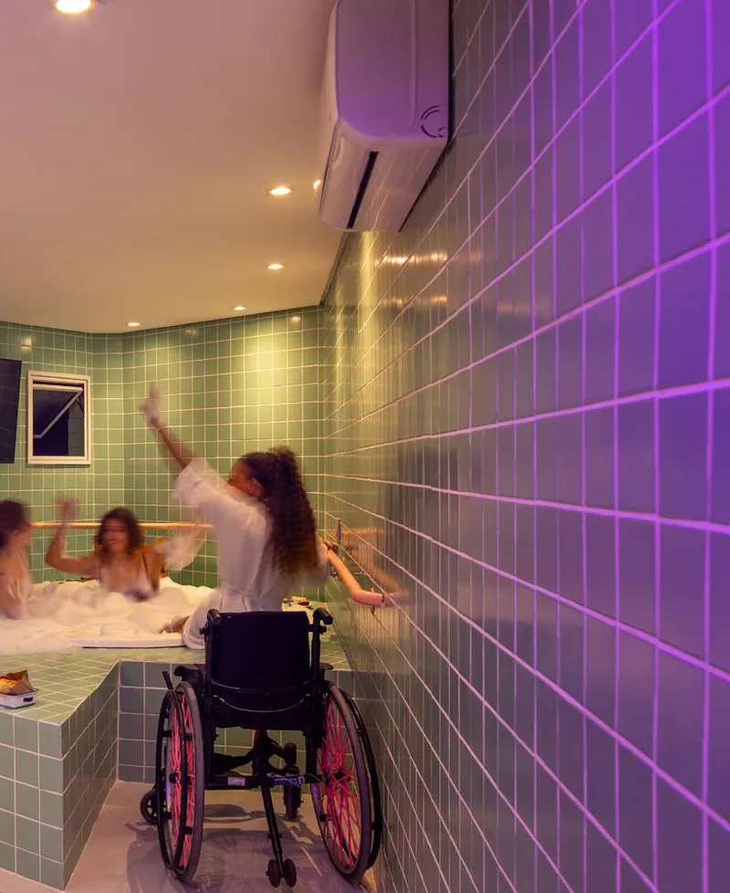 Communal bath space with seated figures and a wheelchair user reaching toward purple-lit tile wall