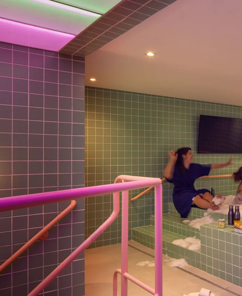 Tiered bathing platform with pink tubular railings against gradient tile walls lit by colored ceiling panels