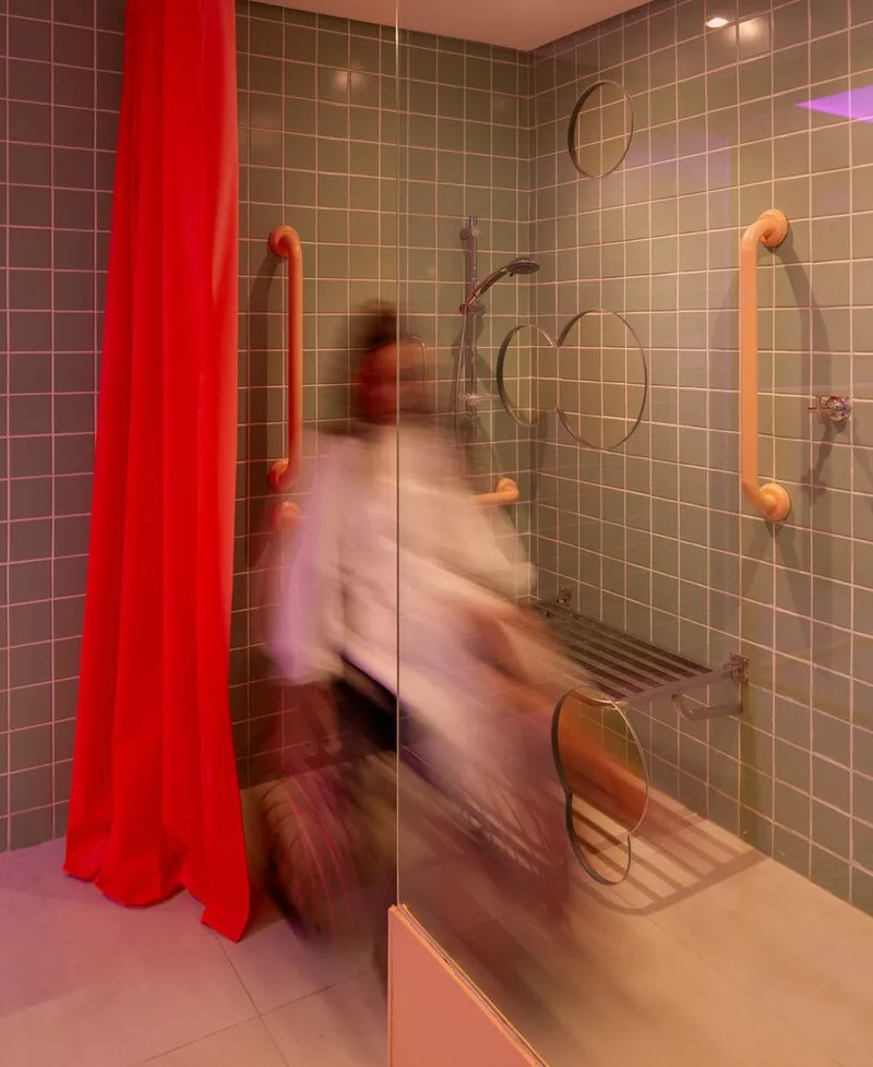 Accessible shower stall with grab bars and handheld showerhead against gradient tile walls illuminated by colored lighting