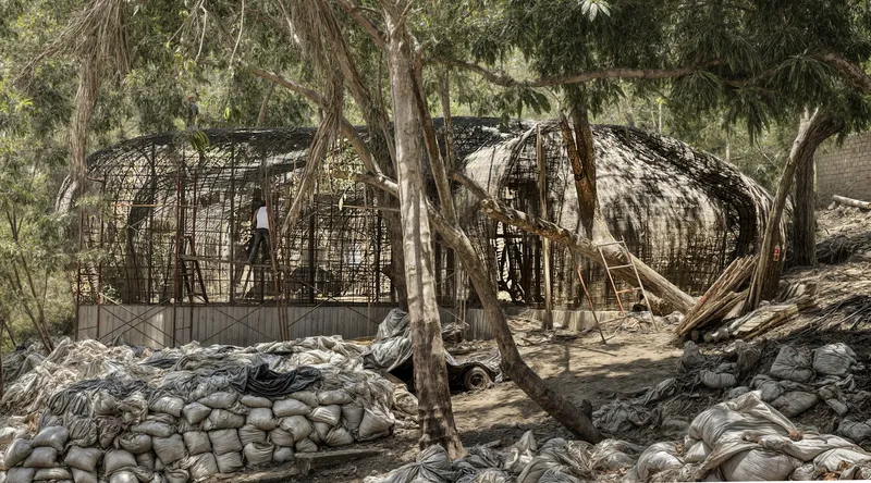 Woven bamboo pavilion structures among trees with sandbags and construction materials in the foreground