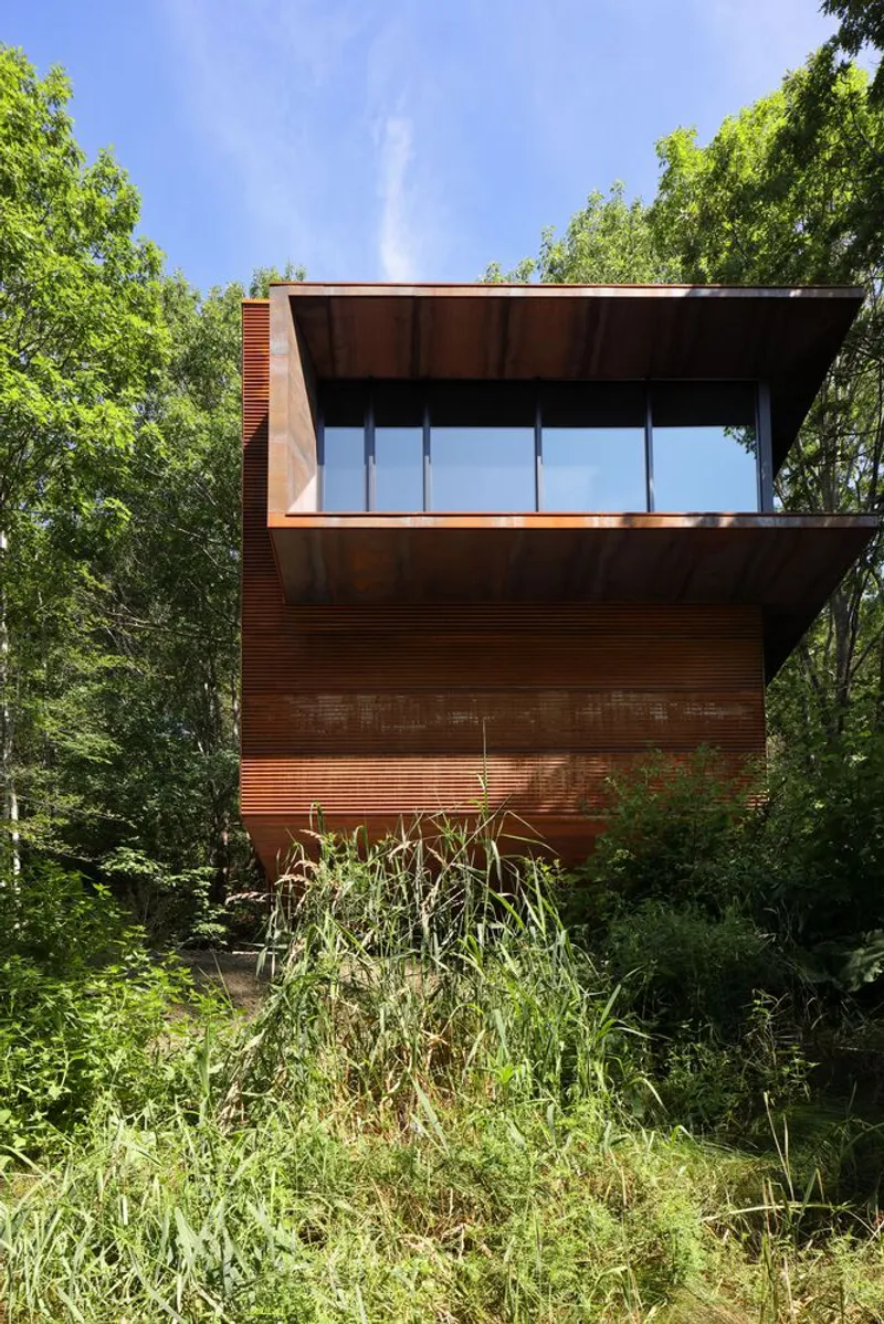 Weathered copper facade with horizontal banding and a ribbon window nestled among dense green trees