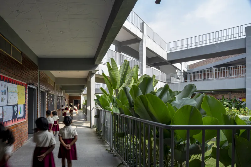 Covered walkway corridor along brick classrooms with students passing planters filled with banana plants and metal railings