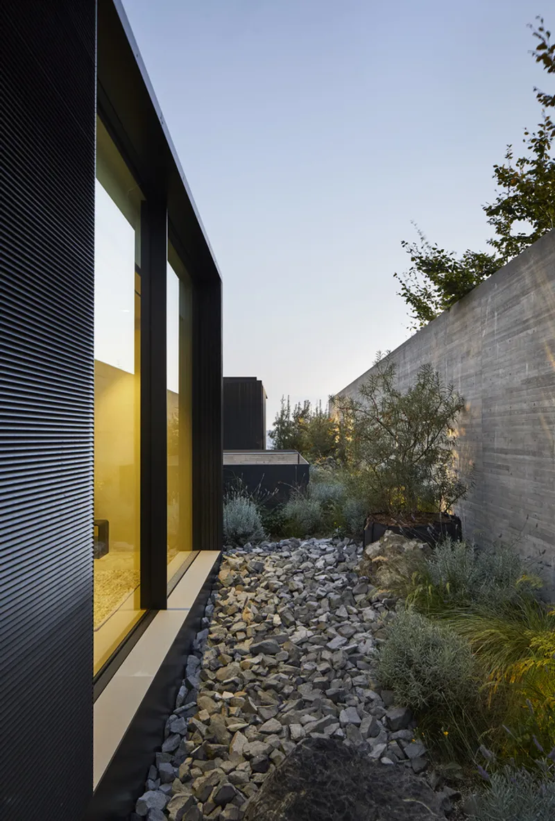 Courtyard path with river rock bed and native grasses between black facade and concrete wall at dusk