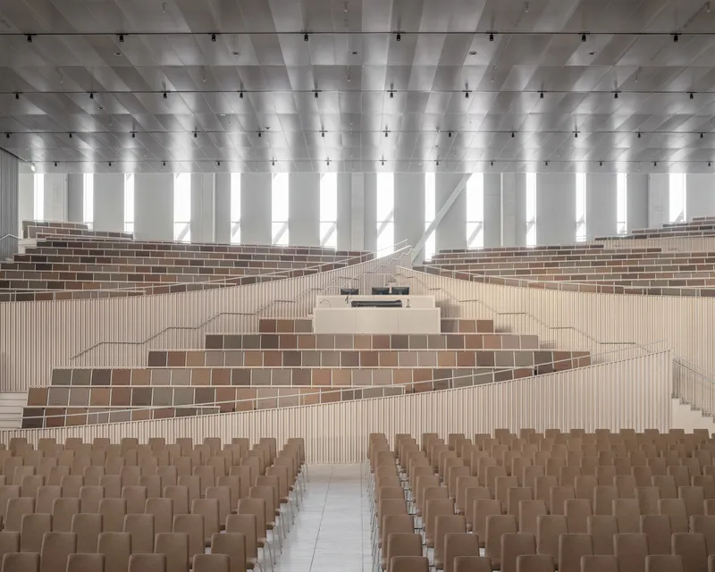 Auditorium interior with tiered timber seating and coffered white ceiling under daylight from tall windows