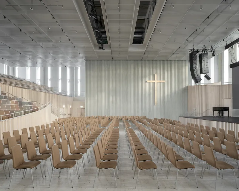 Assembly hall with rows of wood chairs facing a pale wall with cross under a coffered white ceiling