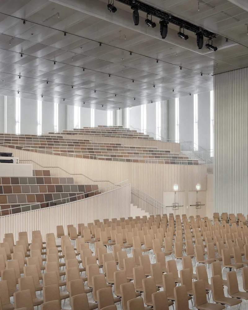 Tiered seating with vertical wood slat fronts and colored tile accents overlooking the lower assembly floor