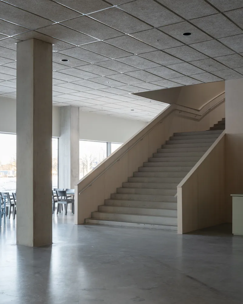 Concrete staircase rising through a lobby with exposed columns and grid ceiling under winter daylight