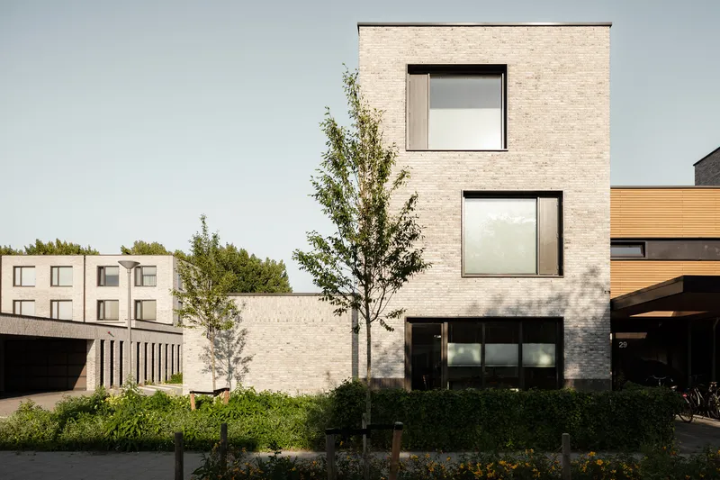 Three-story pale brick volume with stacked windows and a young tree casting shadows on the facade