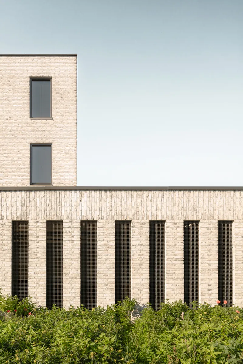 Light brick facade with vertical recessed openings lined with dark mesh above a planted hedge