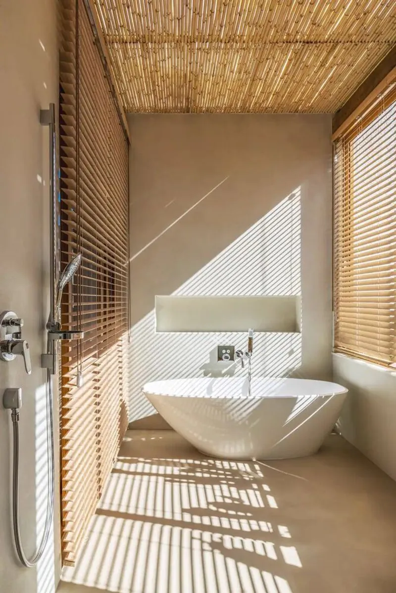 Bathroom with freestanding tub and woven reed ceiling casting striped shadows through timber blinds