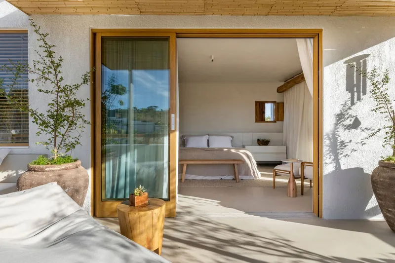 Timber-framed glazed doors opening from terrace to bedroom under wood plank soffit with dappled shadows