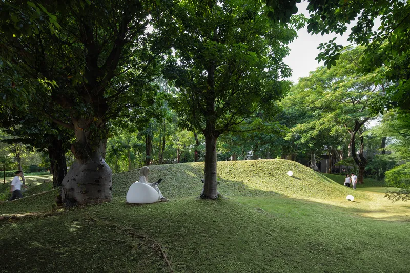 Grass-covered mound roof with white dome skylights emerging between mature trees in a park setting