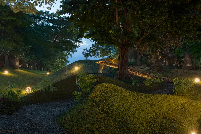 Curved earthen roof structure nestled among trees with pathway lighting at dusk