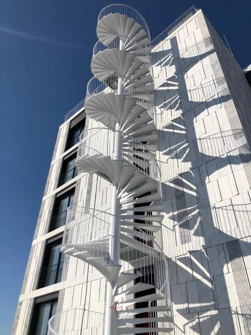 White metal spiral staircase attached to a ribbed facade panel under clear blue sky