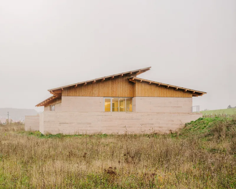 Rammed earth walls with timber cladding and butterfly roof set in a grassy field under overcast sky