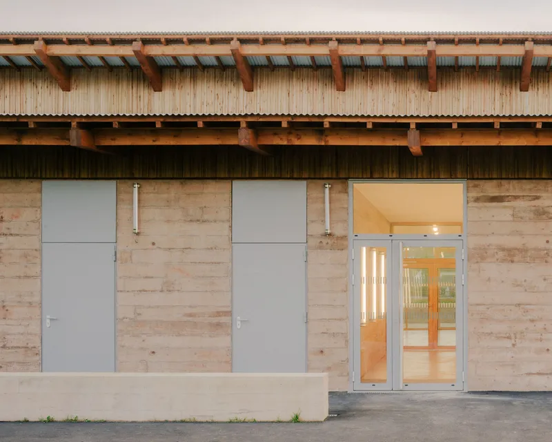 Entrance facade showing rammed earth walls with metal doors and glazed opening below exposed timber eaves