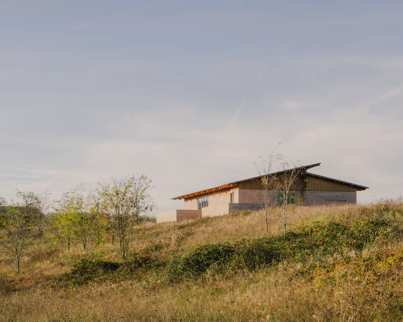 Low-pitched roof volume on a grassy hillside with scattered young trees at dusk