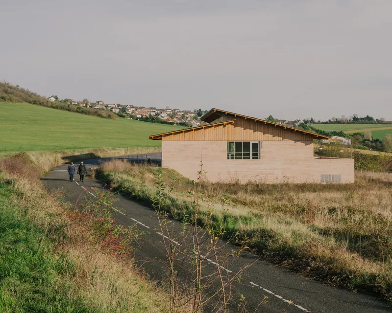 Timber-clad gable structure along a paved path with visitors approaching and farmland beyond