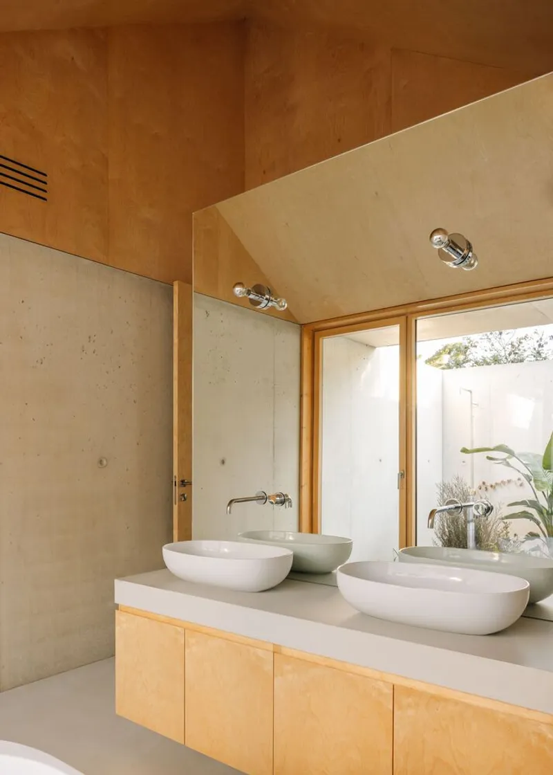 Bathroom vanity with twin vessel sinks, timber cabinetry, and plywood soffit with exposed bulb fixtures looking to planted courtyard
