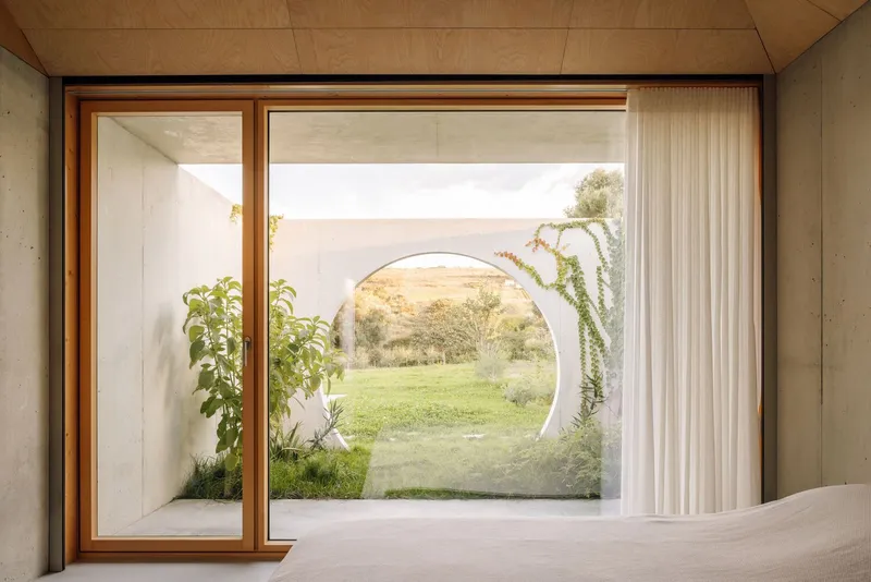 Bedroom view through sliding timber-framed glazing to courtyard with circular moon gate opening onto lawn at dusk