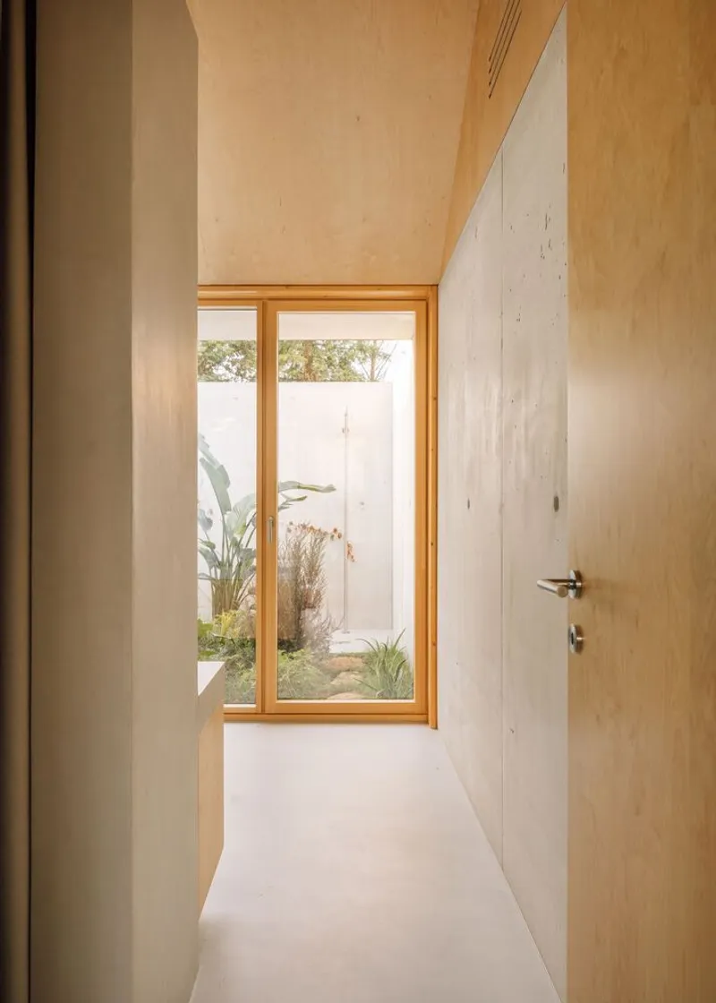 Narrow corridor with plywood doors and ceiling leading to a timber-framed window overlooking a courtyard