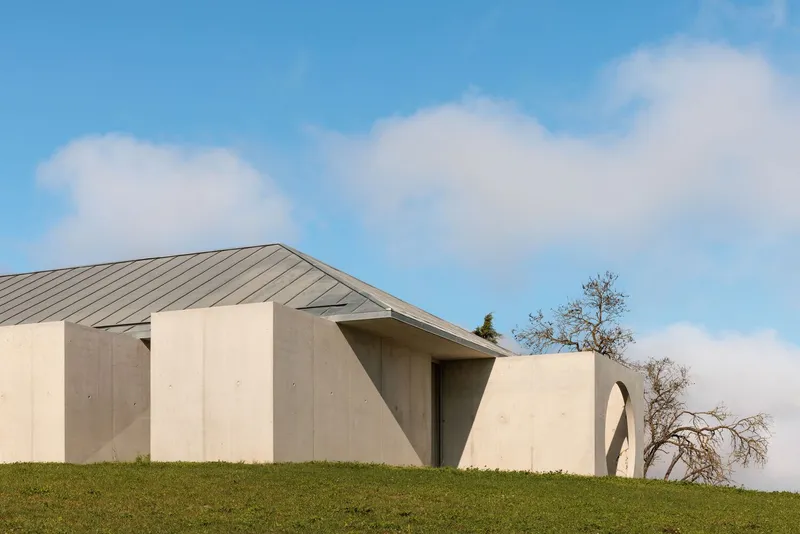 Side elevation showing the angular metal roof meeting concrete walls beside a bare tree