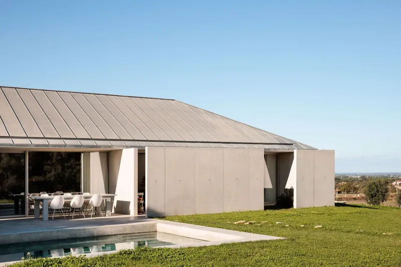 Covered terrace with tapered concrete columns beside a pool and lawn under clear sky