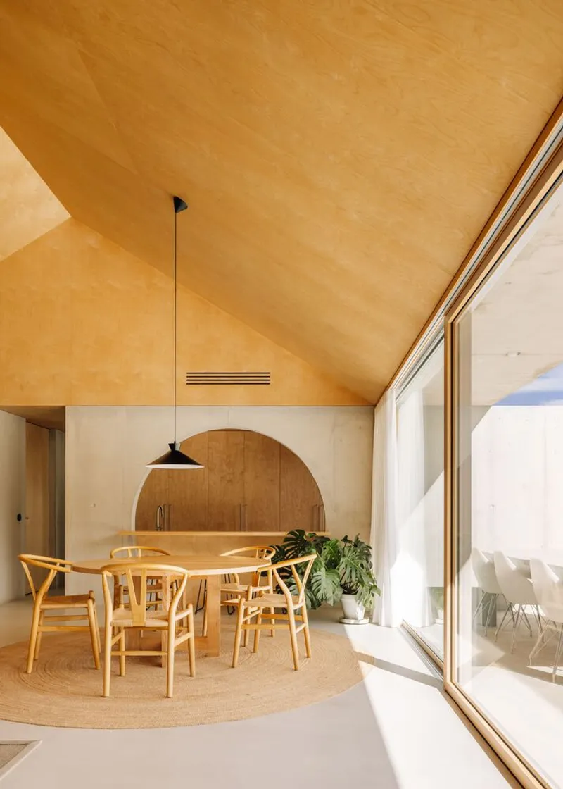 Dining area with plywood vaulted ceiling, arched timber niche, and floor-to-ceiling glazing opening to a courtyard
