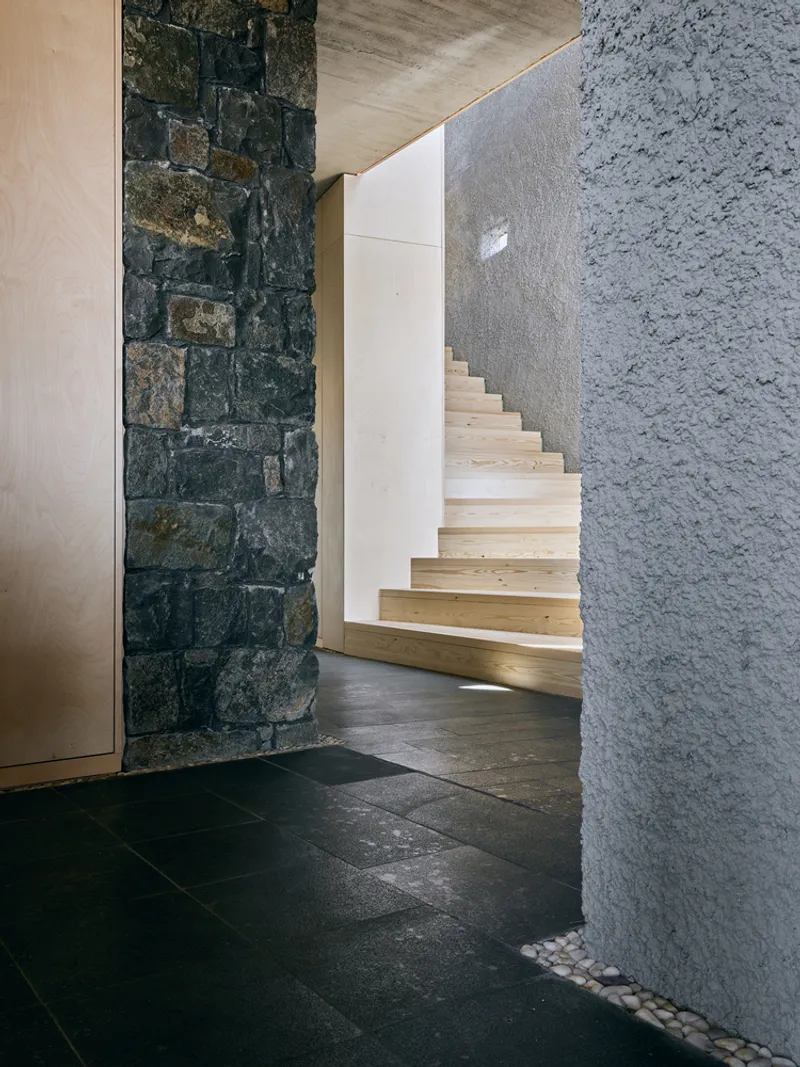 Light wood staircase rising between dark stone wall and pebble-lined textured concrete surface
