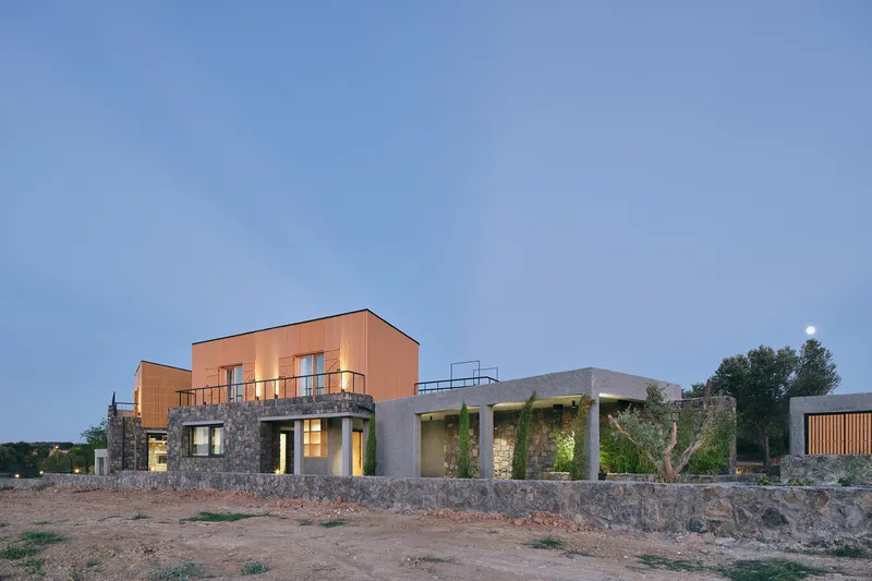 View of the stone and timber volumes with illuminated interiors at dusk