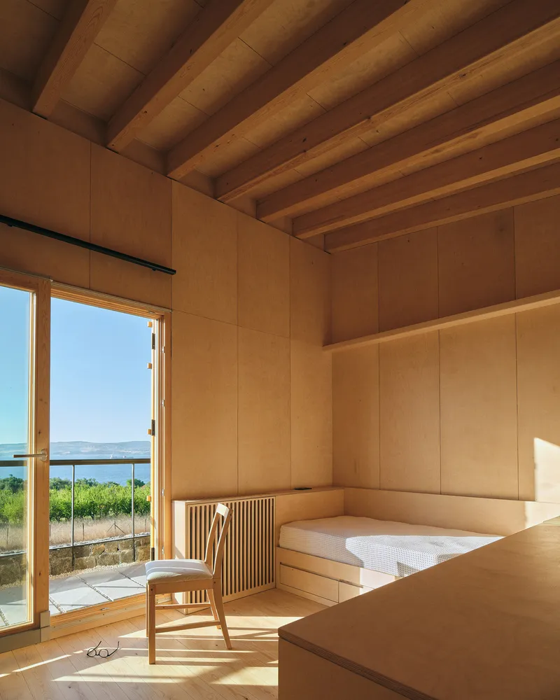 Interior bedroom with exposed timber beams and built-in plywood platform bed overlooking a vineyard and water
