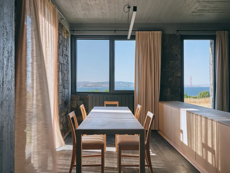 Dining room with stone walls and concrete ceiling framing views of distant hills through tall windows