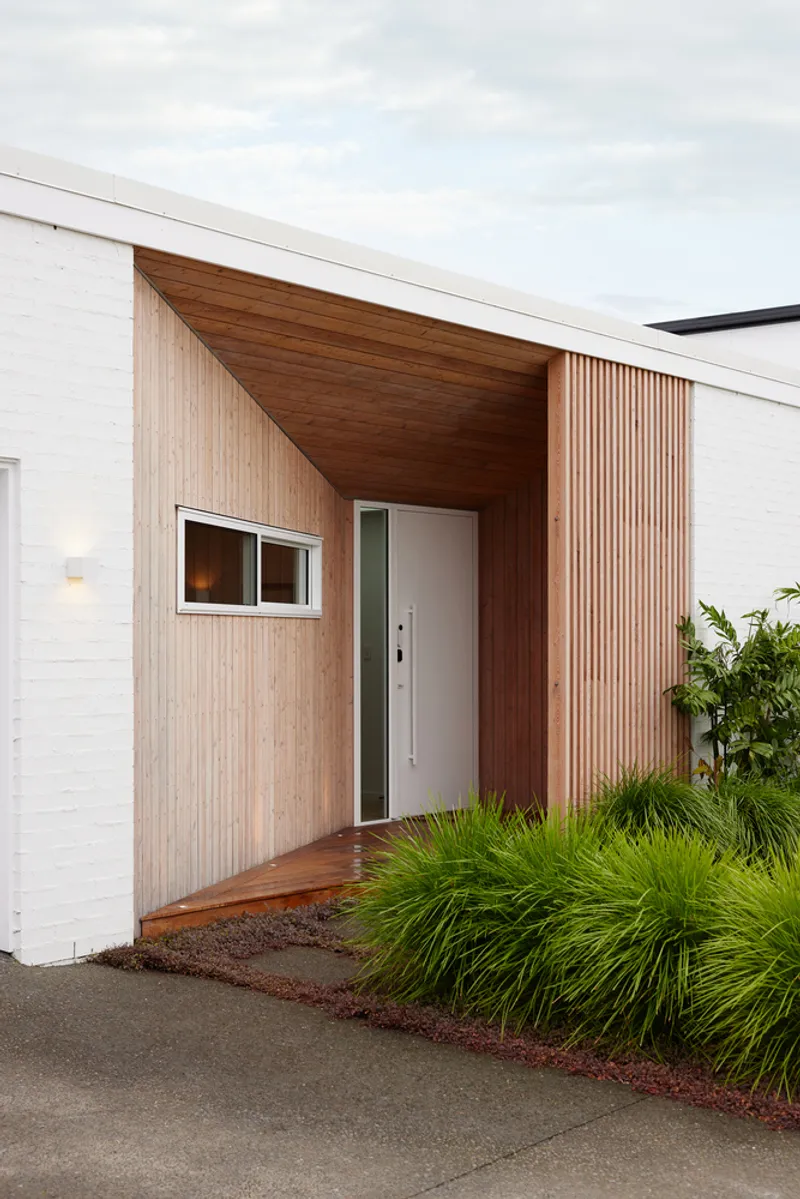 Recessed entry with timber-lined soffit and vertical slat wall beside planted bed with grasses